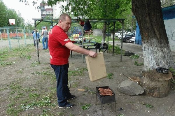 Мужчина во дворе многоквартирного дома раздувает пламя в мангале с шашлыками 